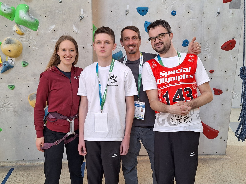 Vor einer Kletterwand stehen vier Personen nebeneinander. Von links nach rechts: Johanna Kolousek (Klettertrainerin), sie trägt einen Klettergurt, Silvio Gorsche, er trägt an einem Schlüsselband einen Ausweis um den Hals, Marco Lamprecht (Klettertrainer), er hält einen Daumen hoch und trägt ebenfalls einen Ausweis an einem Schlüsselband um den Hals und Matthias Zorn, er trägt ein rotes Trikot auf dem in weißen Buchstaben "Special Olympics" steht.