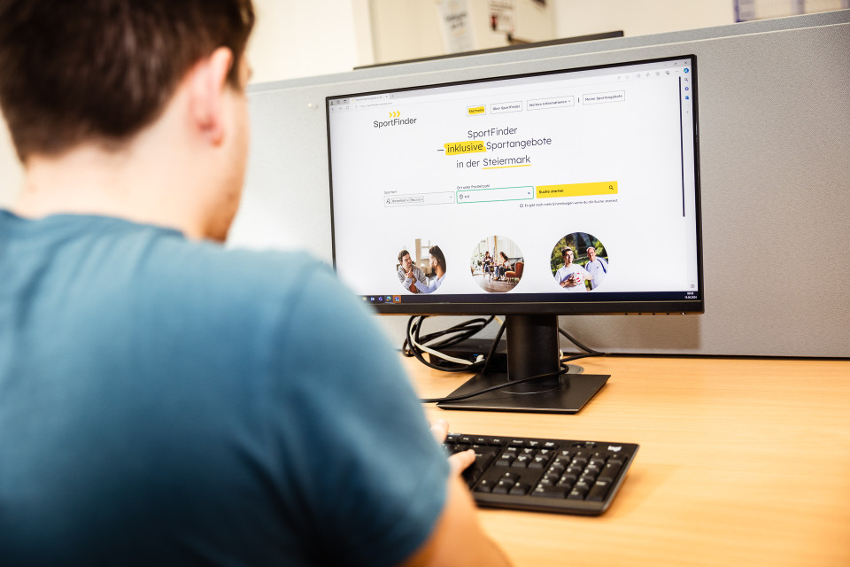 Ein Stand-PC, auf dem die Website des SportFinders aufgerufen ist. Davor sitzt ein Mann in blauem T-Shirt, er ist nur von hinten zu sehen.