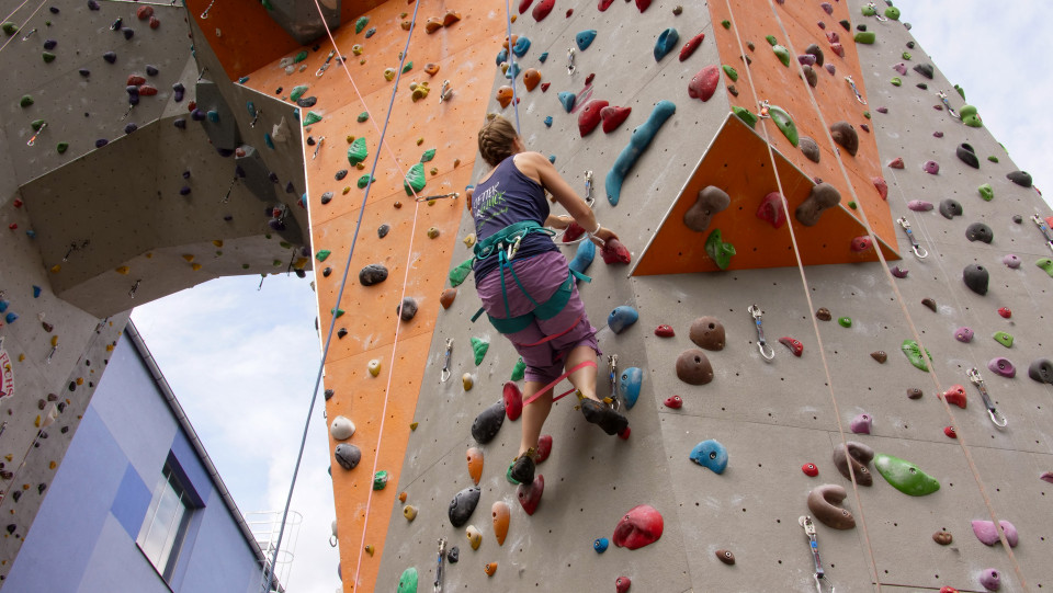 Eine Kletterwand im Freien: Eine Frau klettert, sie ist nur von hinten zu sehen. Um ihre Beine sind zwei rote Gummibänder gewickelt, sodass sie die Beine nicht weit voneinander weg bewegen kann.