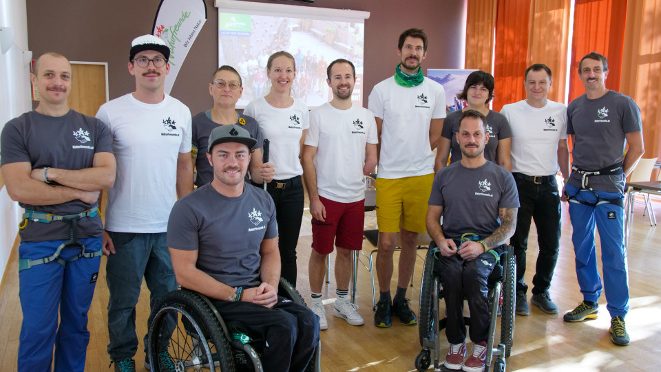 Gruppenfoto von Mitwirkenden des Projekts "Die Kletterwand kennt keine Barrieren". Von links nach rechts, erste Reihe: Angelino Zeller und Markus Pösendorfer (beide sitzen im Rollstuhl). Zweite Reihe: Alexander Guster, Michael Schlegl, Linda Le Bon (sie hält einen Blindenstab in der Hand), Johanna Kolousek, Markus Crell (ihm fehlt der linke Unterarm), Filippo Bignetti, Lisa Marschner, Rene Stix und Marco Lamprecht.