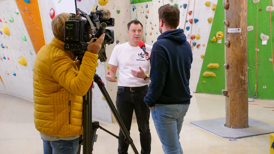 In einer Kletterhalle: Rene Stix, Geschäftsführer der Naturfreunde Steiermark, wird interviewt. Der Interviewer und Kameramann sind nur von hinten zu sehen.
