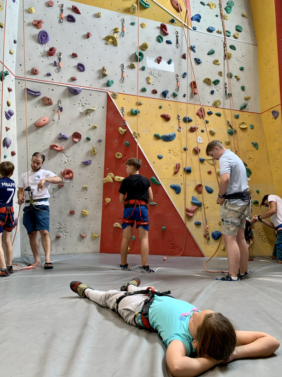 In einer Kletterhalle: Drei Burschen sind von hinten zu sehen, sie tragen Klettergurte an denen bereits Seile befestigt sind, alle stehen aber noch am Boden. Ein Mann mit weißem T-Shirt schaut in Richtung Kamera, auch er trägt einen Klettergurt an dem ein Seil befestigt ist. Im Vordergrund liegt ein Mädchen auf der Klettermatte am Boden, sie trägt ebenfalls einen Klettergurt.