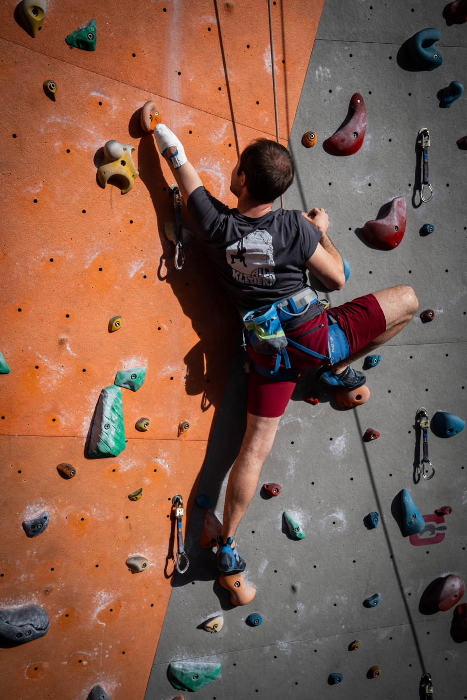 Auf einer halb orangen halb grauen Kletterwand klettert ein Mann. Er ist nur von hinten zu sehen. Seinen linken Arm streckt er zum nächsten Griff aus. Der linke Arm endet knapp unter dem Ellbogen in einem Stumpf, der mit einem Verband umwickelt ist.