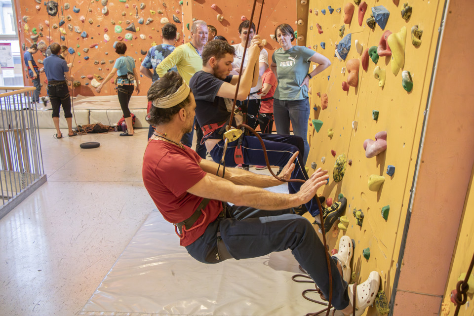 In einer Kletterhalle sind zwei Männer von der Seite zu sehen. Beide hängen im Seil, etwa einen Meter über dem Boden, beide stützen sich mit ihren Füßen an der Wand ab. Der vordere Mann deutet mit beiden Handflächen zur Wand, der hintere umklammert mit beiden Händen das Seil. Im Hintergrund beobachten eine Frau und zwei Männer das Geschehen.