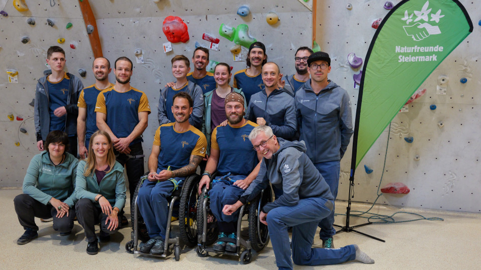 Gruppenfoto von Mitwirkenden und Teilnehmenden des Projekts "Die Kletterwand kennt keine Barrieren" vor einer Kletterwand. Von links nach rechts, erste Reihe: Lisa Marschner, Johanna Kolousek, Markus Pösendorfer, Angelino Zeller (die beiden Herren sitzen im Rollstuhl) und Heinz Pall. Zweite Reihe: Silvio Gorsche, Markus Crell, Ulrich Becker, Serhii Norka, Lisa Butscher, Alexander Guster und Michael Schlegl. Dritte Reihe: Marco Lamprecht, Filippo Bignetti und Matthias Zorn. Rechts neben der Gruppe steht eine grüne Beachflag auf der in weißer Druckschrift die Worte "Naturfreunde Steiermark" zu lesen sind, darüber befindet sich deren Logo in weiß: Drei Blumen über zwei Händen, die einander festhalten.