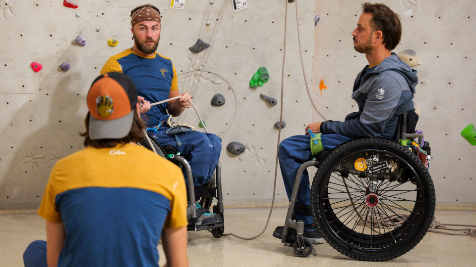 Vor einer Kletterwand sind zwei Rollstuhlfahrer zu sehen, der eine trägt ein gelb-blaues T-Shirt und ein braunes Bandana und sieht in die Kamera. Der andere trägt ine blaue Weste mit weißem Logo am Ärmel und sieht den anderen an. Im Vordergrund ist ein Mann von hinten zu sehen, der im Schneidersitz am Boden Platz genommen hat.
