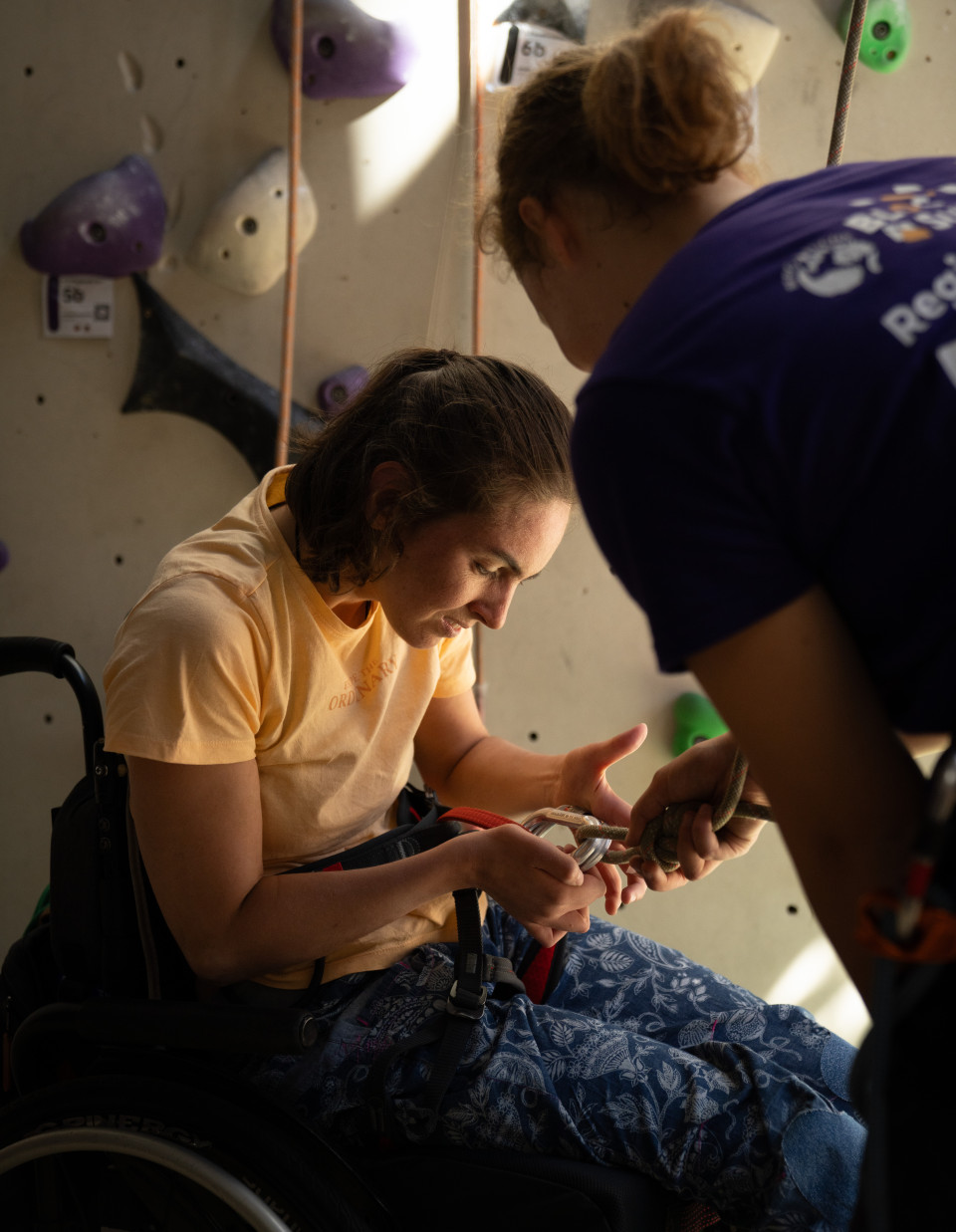 Vor einer Kletterwand: Eine junge Frau im Rollstuhl, trägt einen Klettergurt. Sie sieht sich die Karabiner an, mit denen das Seil am Gurt befestigt ist.