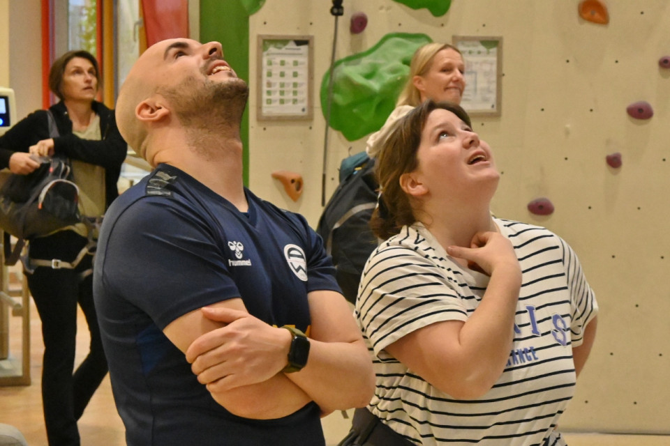 In einer Kletterhalle: Ein Mann mit Glatze und einem blauen T-Shirt sowie eine junge Frau mit schwarz-weiß gestreiftem T-Shirt und Pferdeschwanz stehen nebeneinander und schauen nach oben.