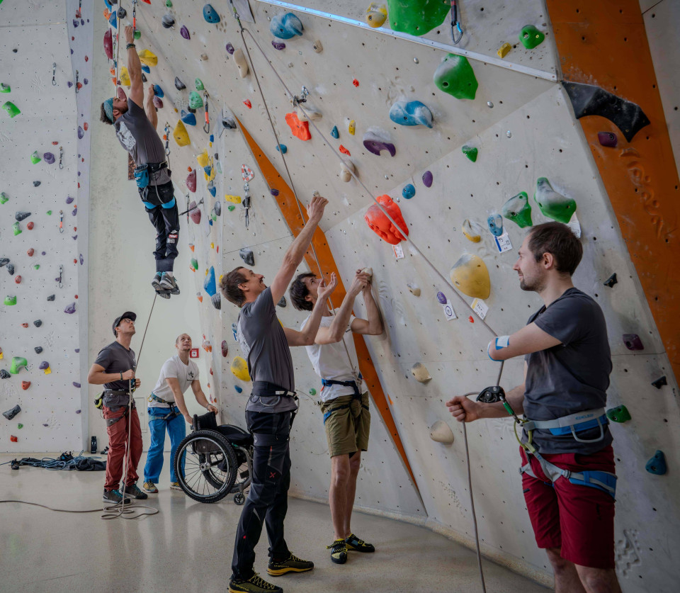 In einer Kletterhalle: Im Vordergrund sind vor einer Kletterwand am Boden stehend drei Männer zu sehen. Zwei davon scheinen sich zu unterhalten und haben den Blick nach oben auf die Kletterwand gerichtet, beide gestikulieren. Einer der beiden wird von dem dritten Mann gesichert. Im Hintergrund sichert ein Mann einen Rollstuhlfahrer, dieser hängt in der Wand. Ein dritter Mann hält am Boden den Rollstuhl bereit.