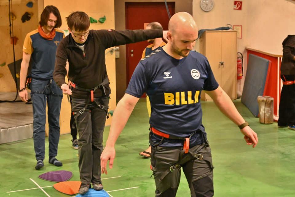 In einer Kletterhalle: Zwei Männer, beide tragen Klettergurte, versuchen auf bunten Pyramidenstümpfen aus Plastik zu balancieren. Der vordere Mann trägt ein dunkelblaues T-Shirt auf dem in großen gelben Blockbuchsteben "Billa" steht. Hinter den zwei Männern steht noch ein dritter, der den Pyramidenstumpf vor sich betrachtet, er trägt ebenfalls einen Klettergurt.